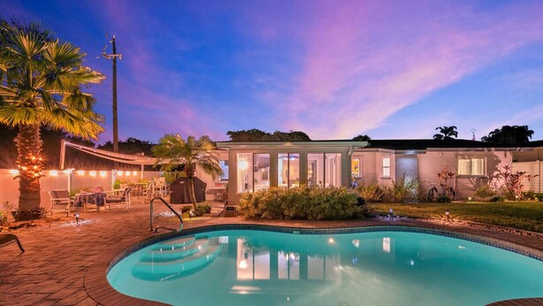 Una piscina con un solarium, una zona relax all'aperto coperta e un edificio con ampie finestre.