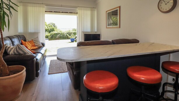 A modern kitchen with a central island, bar stools, pendant lighting, a sofa with cushions, and a view through a glass door. - Rollies Retreat (Fowey)