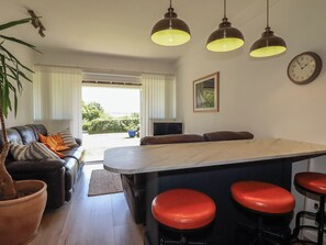 Une cuisine moderne avec un îlot central, des tabourets de bar, des luminaires suspendus, un canapé avec des coussins et une vue à travers une porte vitrée.