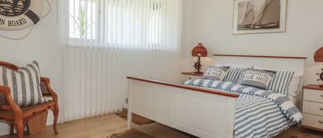 A bedroom with a bed, a nightstand, a lamp, a chair, a window with blinds, a wall clock, and a framed picture.