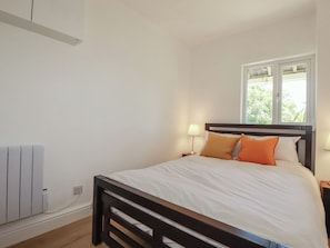 A bedroom with a bed, a window, a nightstand, and a wall-mounted heater. - Rollies Retreat (Fowey)
