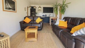 A living room with two leather sofas, a wooden coffee table, a kitchen in the background, and a large plant. - Rollies Retreat (Fowey)