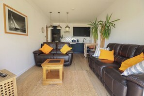 A living room with two leather sofas, a wooden coffee table, a kitchen in the background, and a large plant. - Rollies Retreat (Fowey)