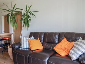 A dark brown leather sofa with striped and solid orange cushions, a wooden dining table with chairs, and two large green plants. - Rollies Retreat (Fowey)