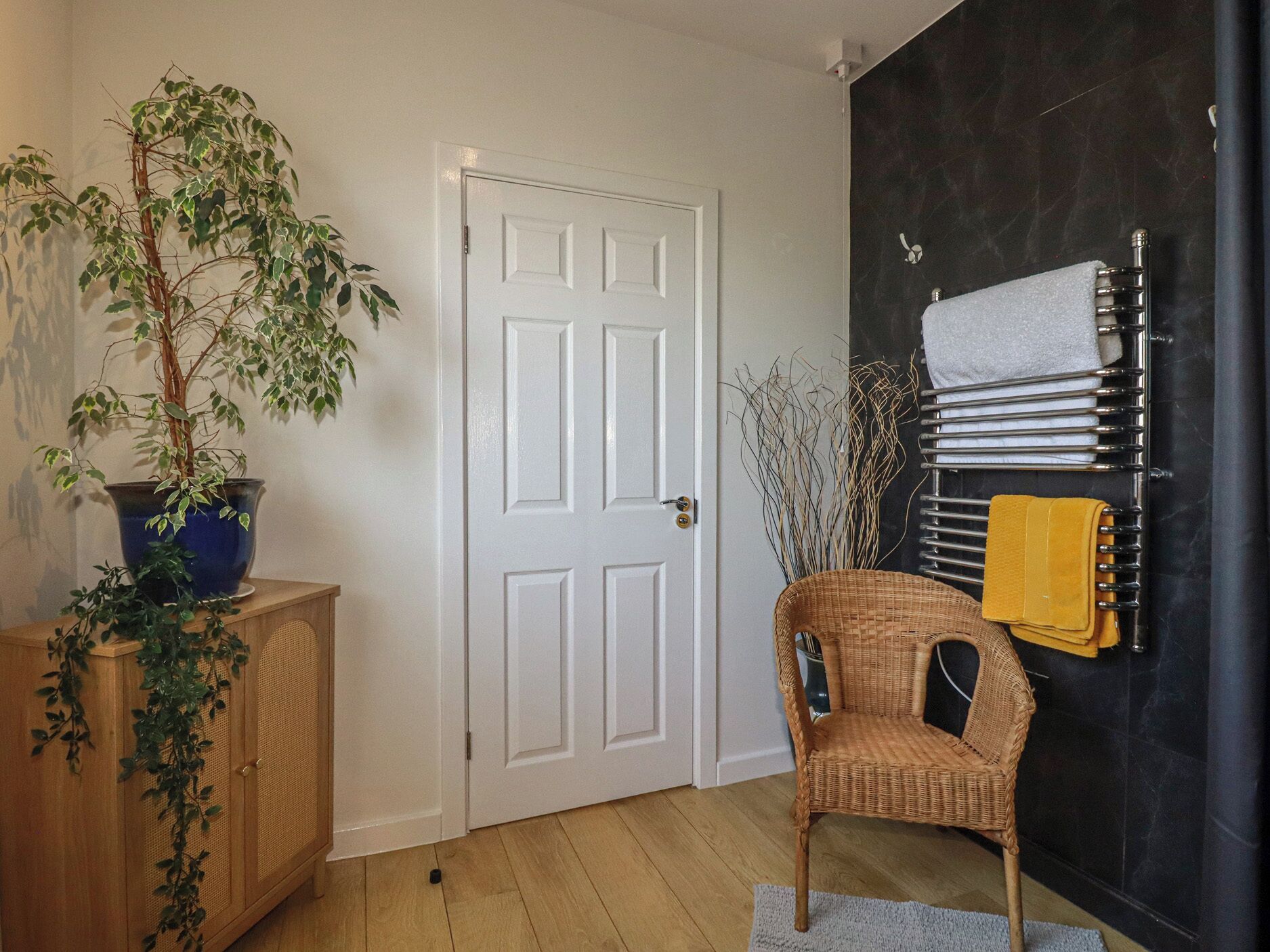 A room with a white door, a potted plant, a wicker chair, and a towel rack with towels.