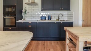 A modern kitchen with dark cabinetry, a built-in oven, a microwave, a stainless steel sink, and a wooden dining table. - Rollies Retreat (Fowey)