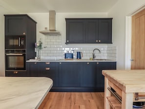 A modern kitchen with dark cabinetry, a built-in oven, a microwave, a stainless steel sink, and a wooden dining table. - Rollies Retreat (Fowey)