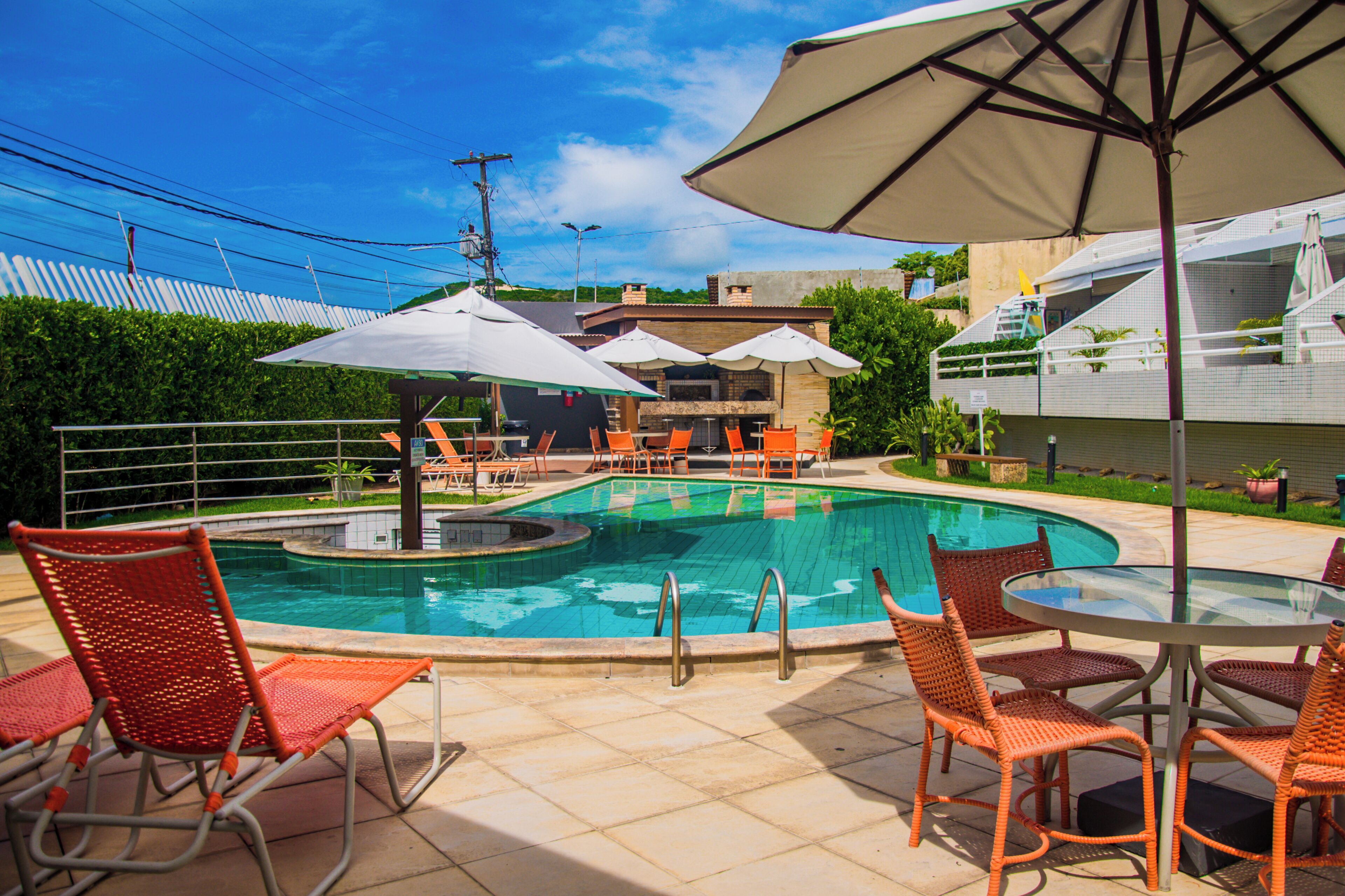 A swimming pool with lounge chairs and umbrellas, surrounded by a fence and buildings.
