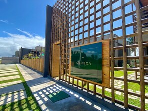 A modern building with a wooden lattice facade, a green sign with 'GAMBOA' text, and a paved pathway with grass. - TOCA GAMBOA - Apartment 190m from Patacho Beach on the Ecological Route of Miracles - AL (Porto de Pedras)