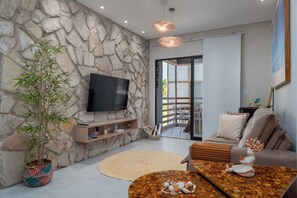 A living room with a stone wall, a flat-screen TV, a sofa, a coffee table, and a potted plant. - TOCA GAMBOA - Apartment 190m from Patacho Beach on the Ecological Route of Miracles - AL (Porto de Pedras)