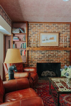 Ein gemütlicher Wohnraum mit Kamin aus Backstein, einer Ledersofa, einem Bücherregal und einem gerahmten Bild über dem Kamin.