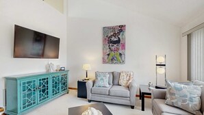 Un salon comprenant un canapé, une table basse, un meuble de couleur turquoise, un téléviseur à écran plat et un tableau accroché au mur.
