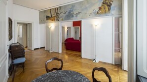 Ein geräumiges Zimmer mit Holzboden, einem Esstisch mit Stühlen, einem roten Sofa und einem großen Wandbild mit Tieren und Vögeln.