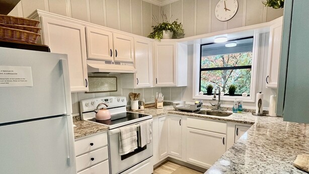 A kitchen with white cabinets, a stove, a refrigerator, a window with a view of greenery, and a clock on the wall. - The Sanctuary in Warm Springs GA (Warm Springs)