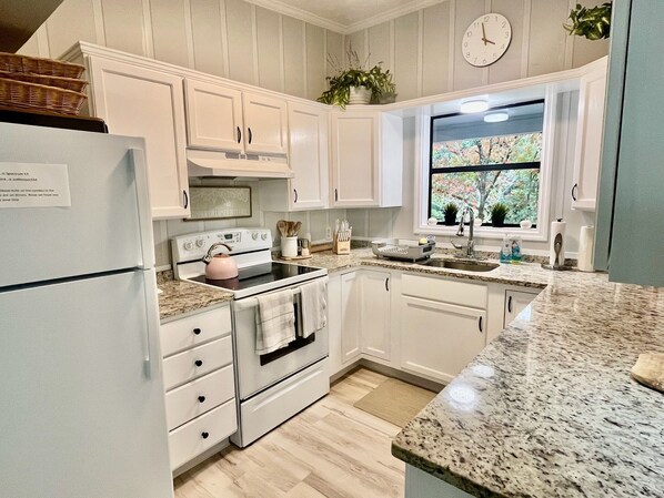 Una cocina con armarios blancos, estufa, refrigerador, ventana con vista a follaje y un reloj en la pared.