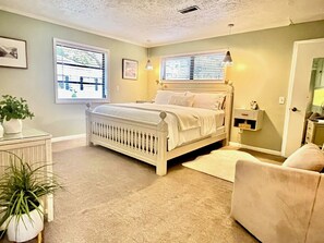 Un dormitorio con una cama blanca, una mesita de noche, una silla, una pequeña mesita con lámpara y una ventana con persianas.