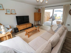 A living room with a sofa, coffee table, TV, and a sliding glass door leading to an outdoor area. - 6 Chapel Rock Close (Perranporth)