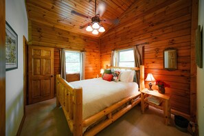 Quarto de cabana de madeira com cama, mesa de cabeceira, abajur e ventilador de teto.