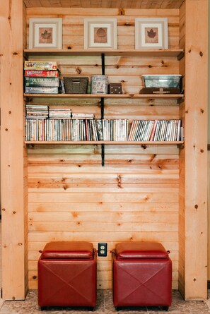 Quarto de madeira com estantes contendo livros e jogos, dois pufes de couro vermelho e quadros nas paredes.