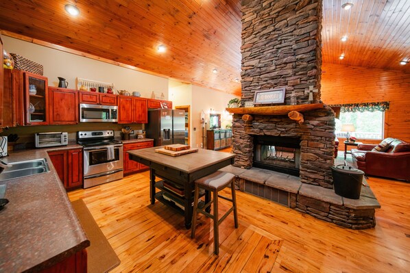 A spacious kitchen with wooden cabinets, a stone fireplace, and a dining area with a bench. - Big Rock | Theater Room, Hot Tub, Hocking Hills (Rockbridge)