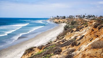 Un paisaje costero con una playa arenosa, acantilados rocosos y una costa distante con edificios.