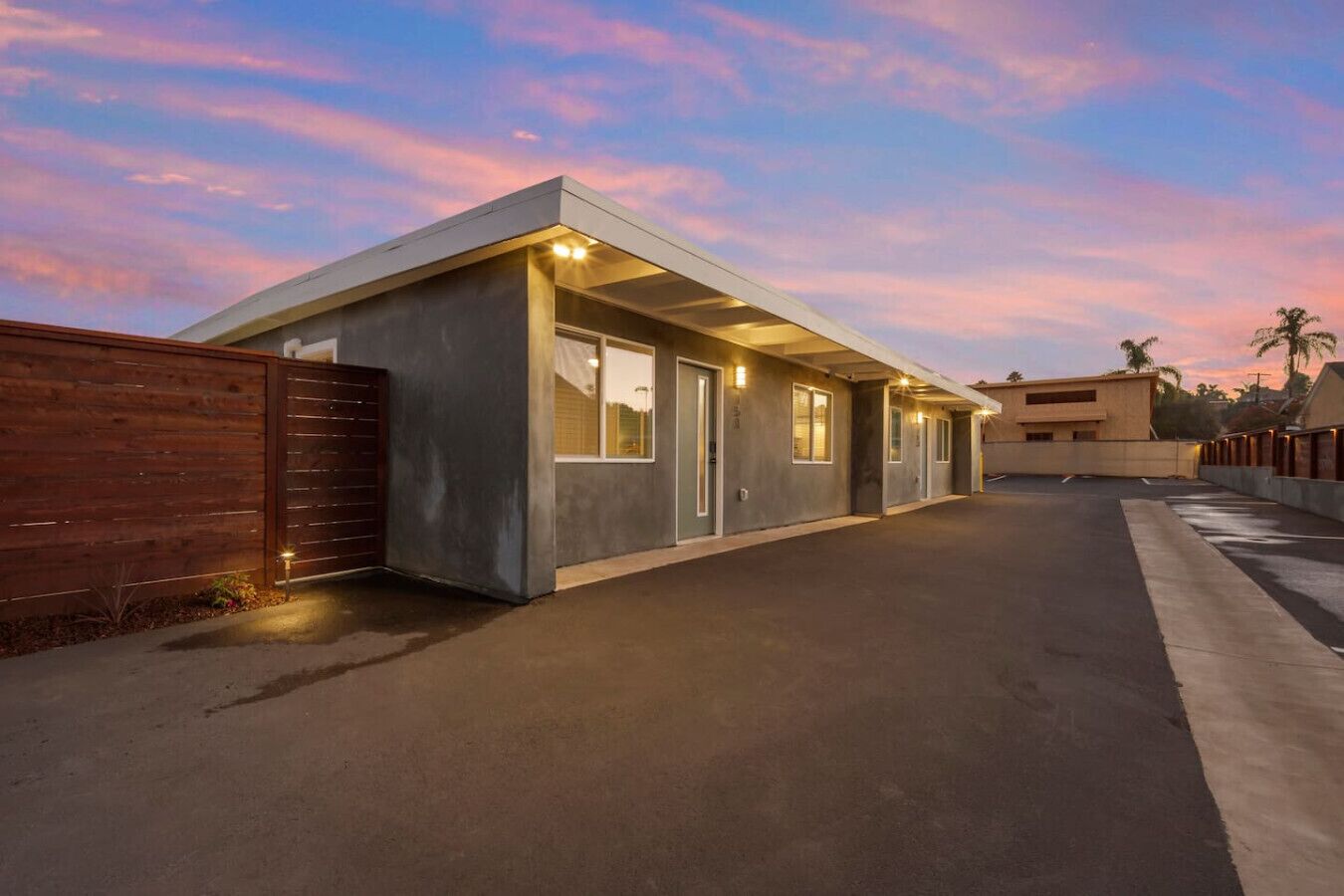 A modern building with a flat roof and large windows, surrounded by a wooden fence and a paved driveway.