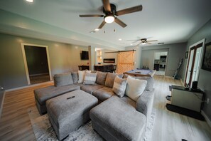 A modern living room with a sectional sofa, a ceiling fan, and a kitchen in the background. - Whispering Acres | Hocking Hills, Hot Tub, Firepit (Logan)