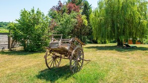 Ein antiker Holzwagen auf einer Wiese mit Bäumen und einem Holtagebau im Hintergrund.