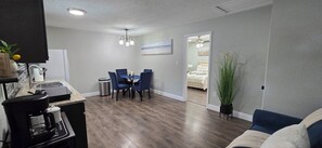 Una cocina moderna con cafetera, un comedor con sillas azules, piso de madera y un espejo grande que refleja un dormitorio.