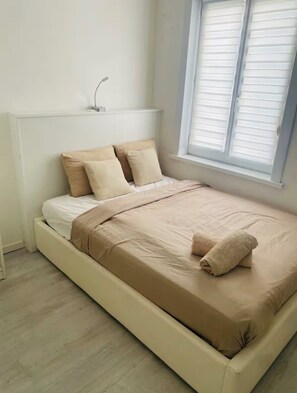 Un letto singolo con materasso e cuscini color beige, un comodino con una lampada e una finestra con tende.