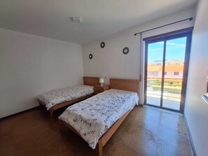 Una habitación con dos camas individuales, una mesita de noche pequeña con una lámpara, una luz en el techo y un ventanal corredero que da a un espacio al aire libre.