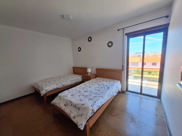 A room with two single beds, a small bedside table with a lamp, a ceiling light, and a sliding glass door leading to an outdoor area.