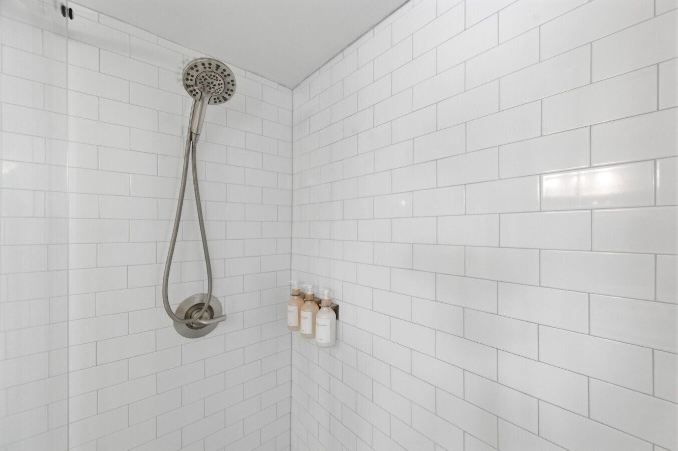 Une douche équipée d’un pommeau de douche à main et d’un robinet mural, des murs recouverts de carreaux blancs et une étagère pour les articles de toilette.