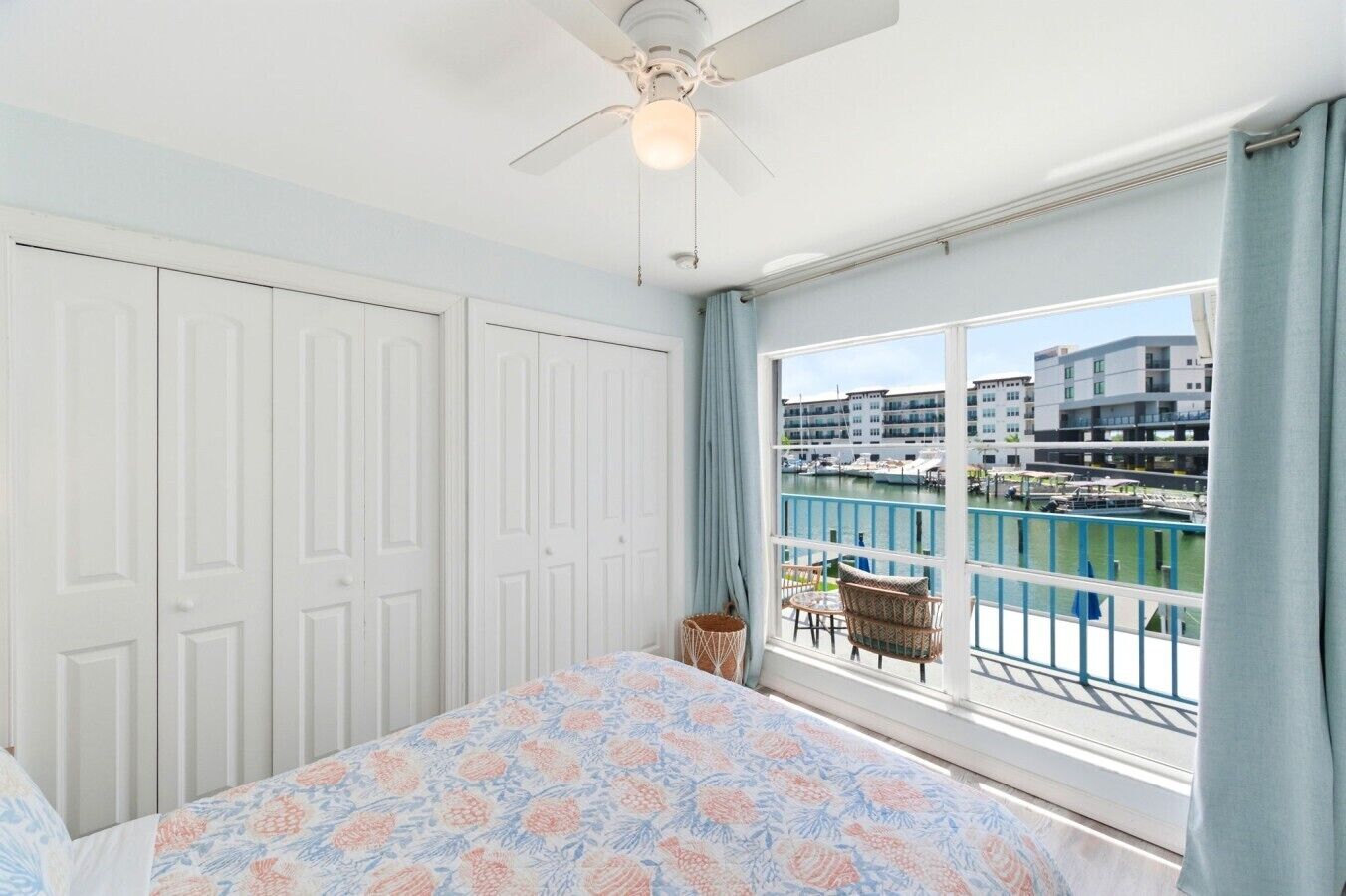 A bedroom with a bed, a ceiling fan, a window with a view of a marina, and a balcony with wicker furniture.