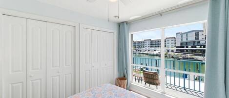 A bedroom with a bed, a ceiling fan, a window with a view of a marina, and a balcony with wicker furniture.