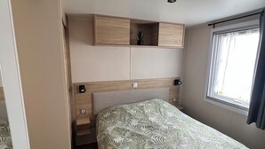 A bedroom with a bed, a headboard, a window with curtains, and a built-in shelf. - Mobil-Home 417 6 persons (Litteau)