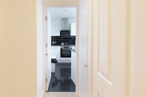 A narrow hallway leading to a kitchen with white cabinets, a black tiled backsplash, and a stainless steel range hood. - 3BR Flat in Hyde Park Area, Steps Away from Oxford Street (London)