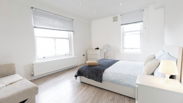 Ein Schlafzimmer mit Bett, Nachttisch, Lampe und einem Fenster mit Jalousien.