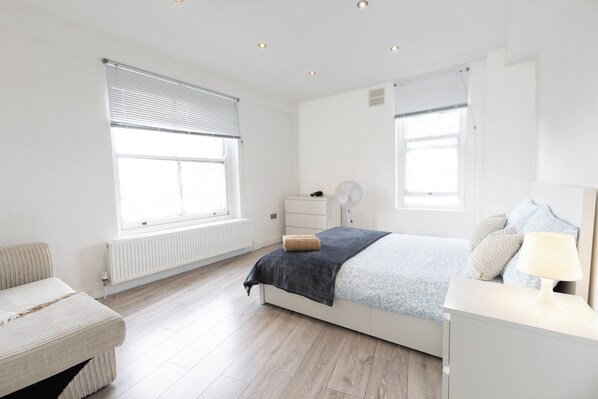 A bedroom with a bed, bedside table, lamp, and a window with blinds. - 3BR Flat in Hyde Park Area, Steps Away from Oxford Street (London)