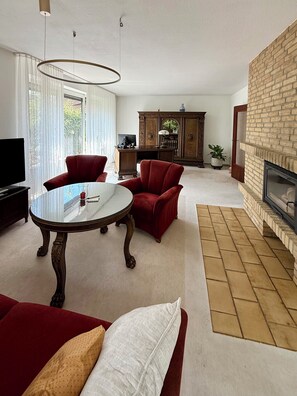 Een woonkamer met een open haard, een televisie, een eettafel en rode fauteuils.