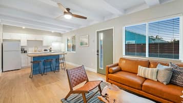 A modern kitchen with white cabinets, a central island with stools, a ceiling fan, and a large window with blinds.