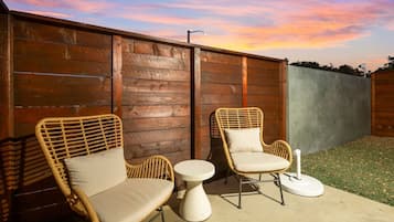 A patio with two wicker chairs and a small round table, set against a wooden fence with a sunset in the background.