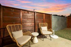 An outdoor seating area with wicker chairs and a small round table, set against a wooden fence with a sunset in the background. - Downtown Haven|Patio|Near Dining (Vista)