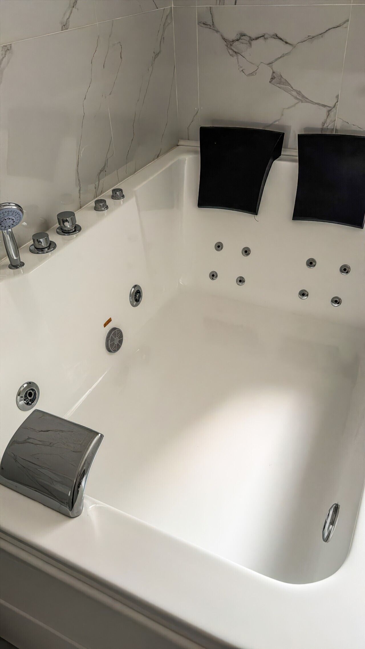 A white bathtub with multiple faucets and a drain, set against a marble wall.