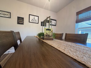 Een eetgedeelte met een houten tafel, stoelen, afbeeldingen in lijsten aan de muur en een raam met jaloezieën.