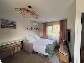 A bedroom with a bed, a desk, two chairs, a ceiling fan, a TV, and a window with curtains.