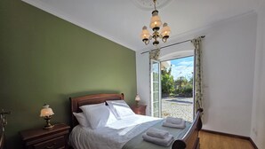 Ein Schlafzimmer mit einem hölzernen Bett, einem Nachttisch mit Lampe, einem Fenster mit Vorhängen und Blick ins Freie.