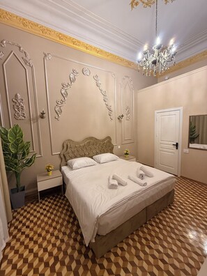 Quarto com piso quadriculado, cama com roupa de cama branca, lustre e molduras decorativas nas paredes.