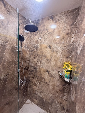 A shower with a handheld showerhead and a glass-enclosed rain shower head, a marble wall, and a shelf with toiletries. - Crown Palace Hotel Baku (Baku)