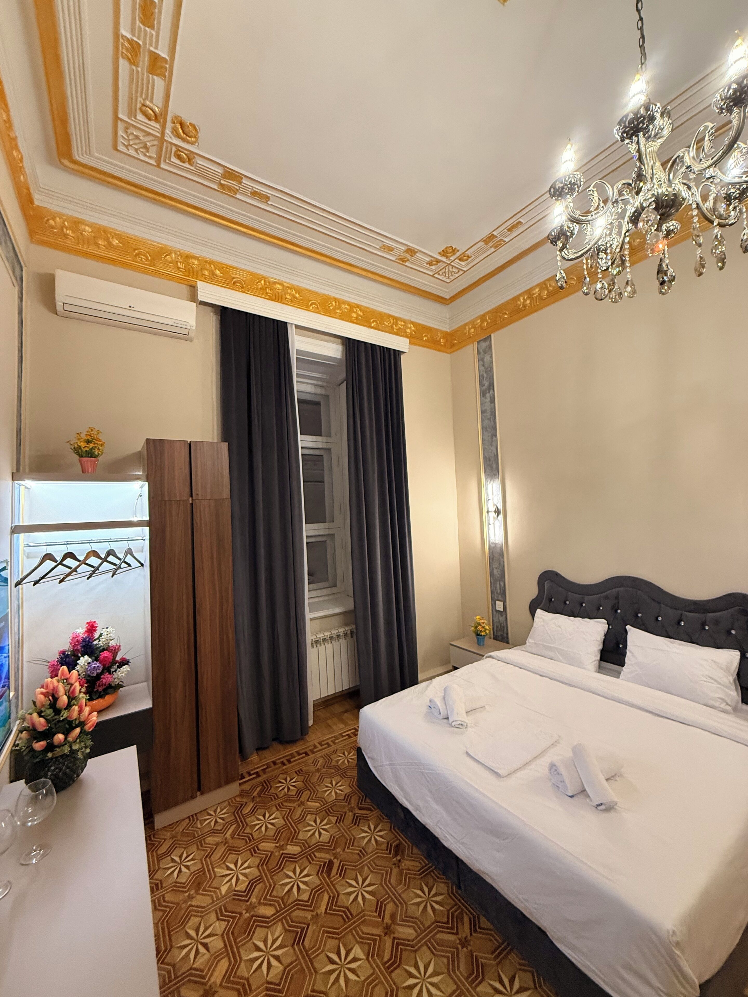 A hotel room with a bed, a chandelier, a dresser, a mirror, and a window with curtains.